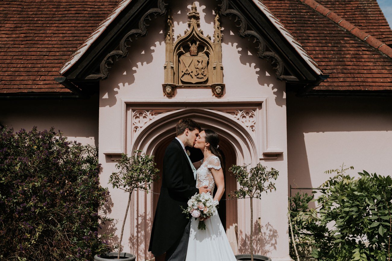 Bride and Groom stand in front of gothic, pink, gatekeepers lodge
