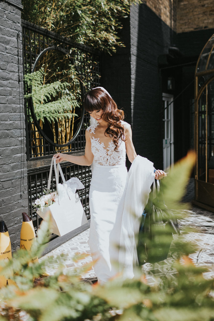 Brunswick House Wedding: Bride carrying bags in a courtyard, wearing a white lace gown and veil, with caution signs nearby.