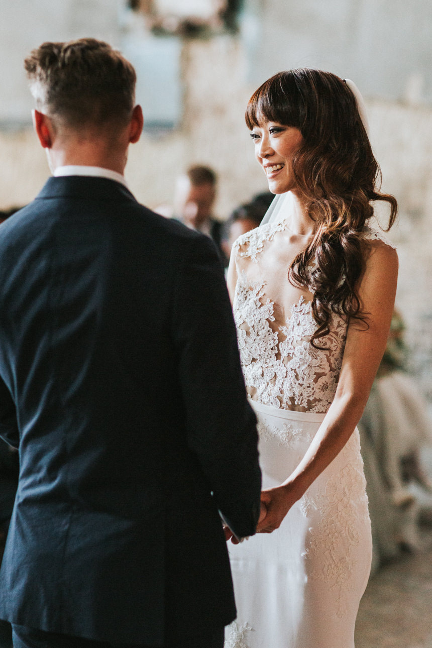Bride smiles at her groom during their Brunswick House and Asylum Chapel Wedding ceremony. She wears a white lace gown with a sheer neckline and her hair is styled in loose waves.