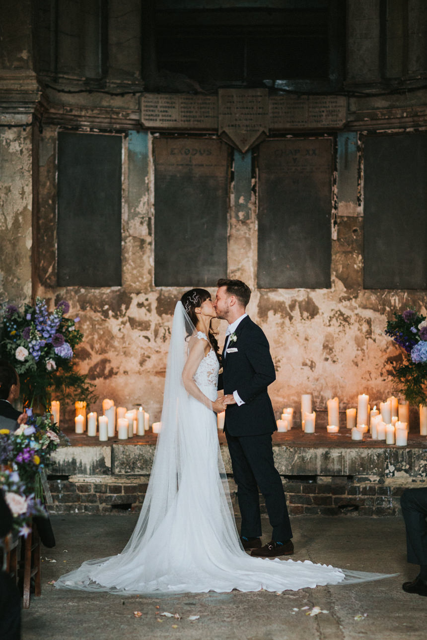 A bride and groom share a kiss during their Wedding ceremony. Candles and flowers decorate the altar in the historic venue.