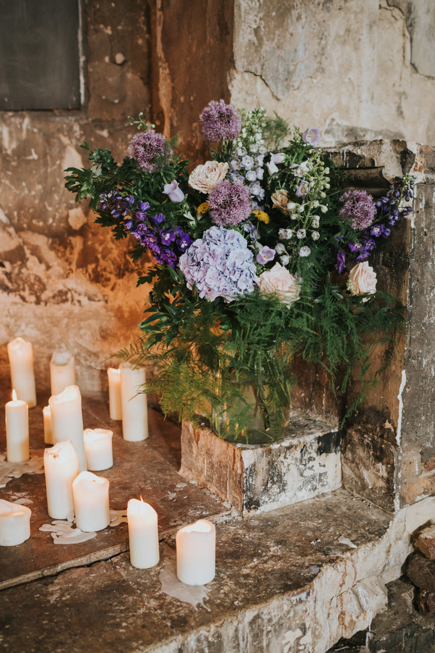 A lush floral arrangement of purples, pinks, and greens sits in a glass vase on a stone ledge, surrounded by lit candles. Romantic Brunswick House wedding decor.