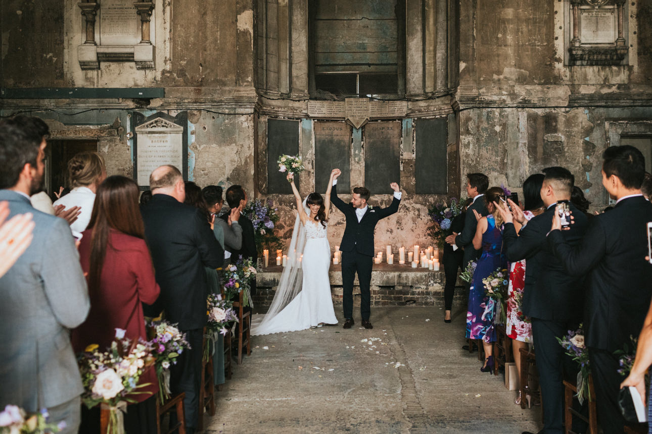Bride and groom celebrate their wedding, arms raised in victory, walking down the aisle lined with cheering guests and candlelight.