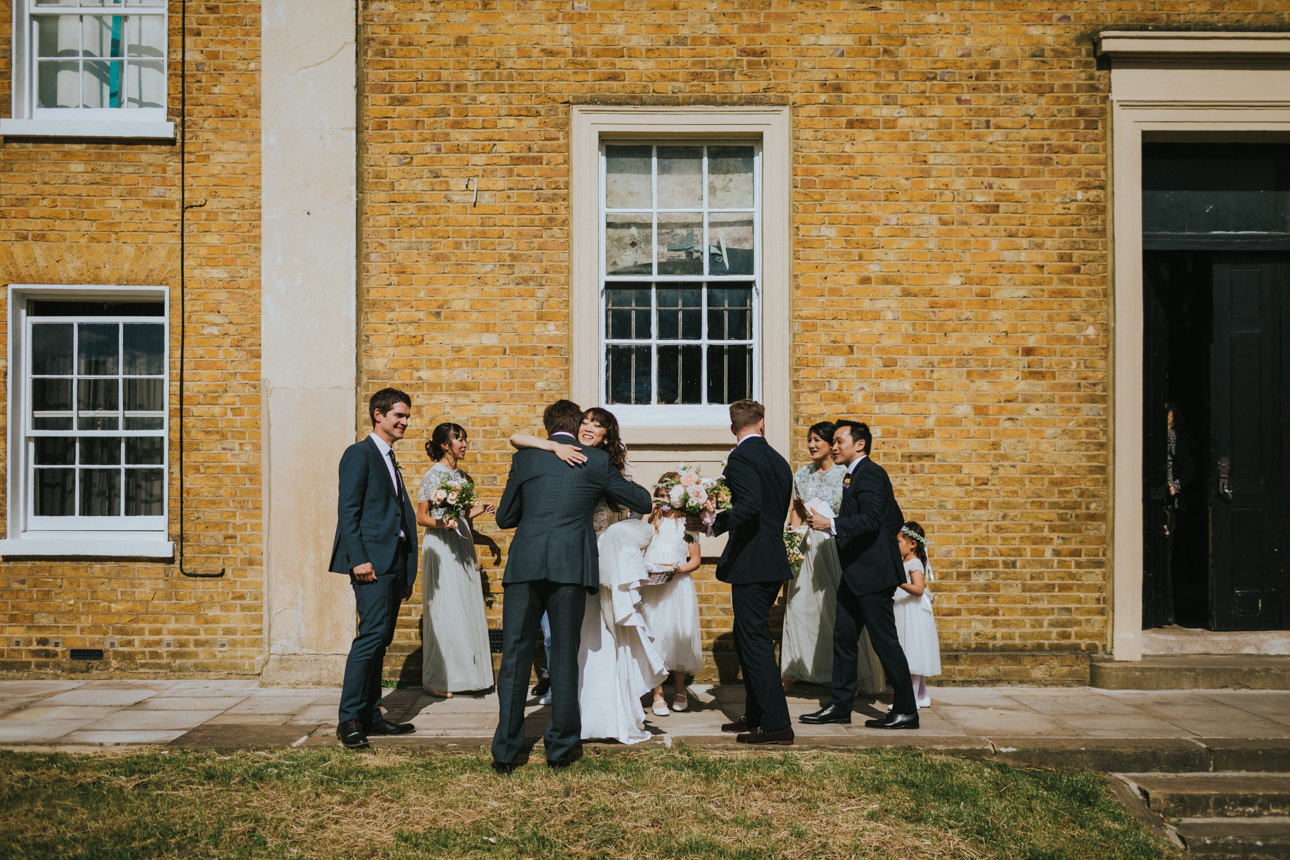 A Brunswick House Wedding scene: The wedding party gathers outside a yellow brick building, the bride hugging a guest, bridesmaids in light dresses, and groomsmen in dark suits.