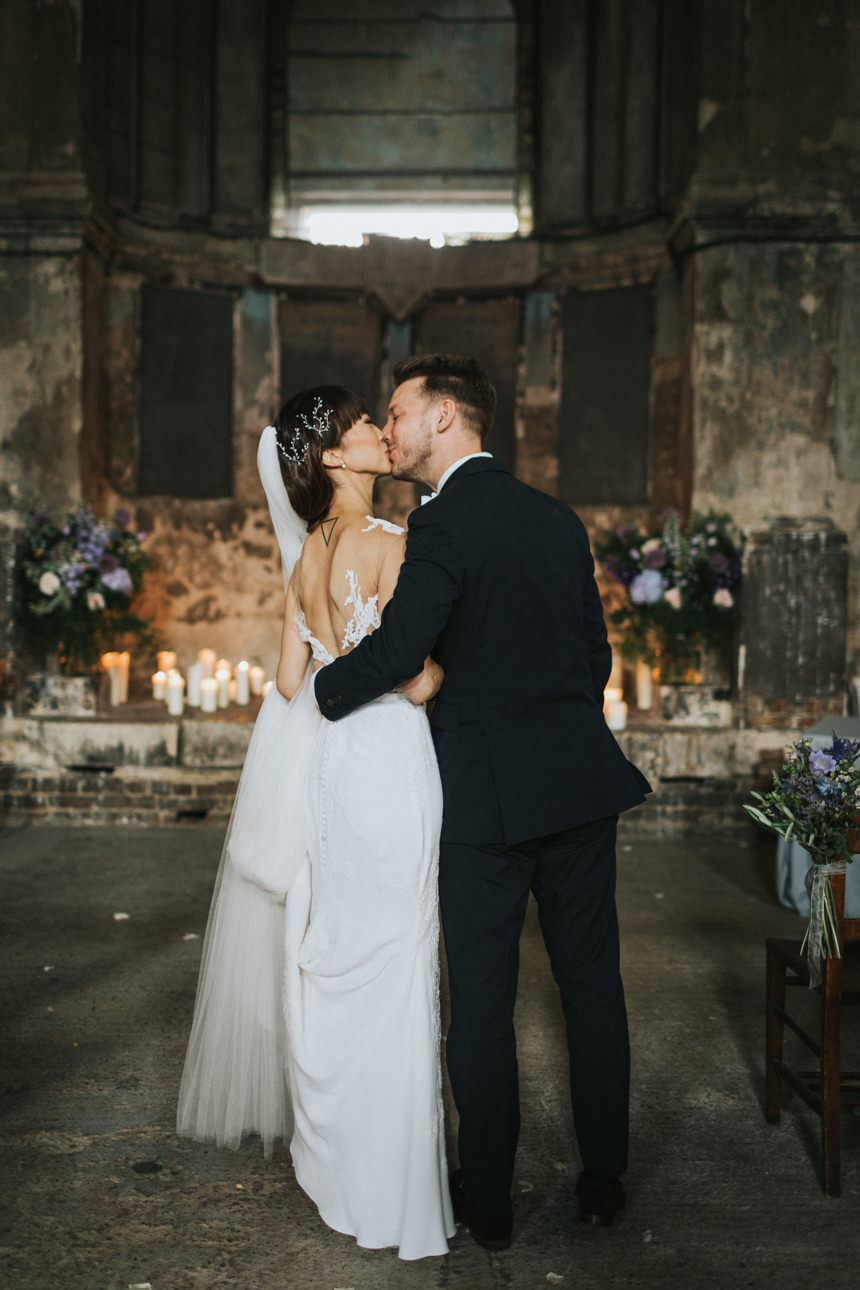A bride and groom share a kiss at their Brunswick House and Asylum Chapel Wedding. She wears a white dress with lace details, and he wears a black tuxedo. Candles and floral arrangements decorate the historic venue.