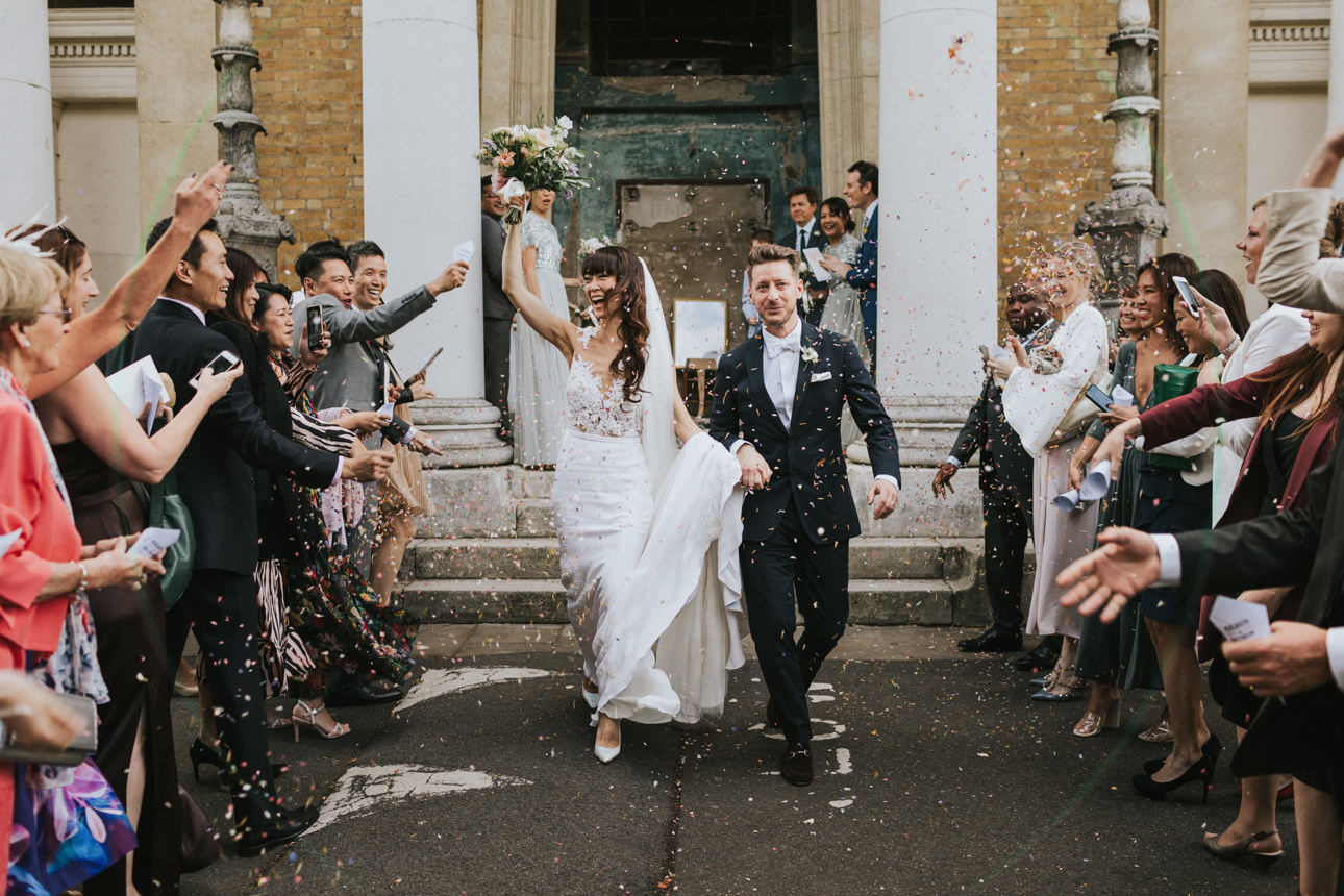 A joyful Brunswick House and Asylum Chapel Wedding scene: a bride and groom exit the venue amidst a shower of confetti and cheering guests. The bride, in a white gown, raises her bouquet in celebration.