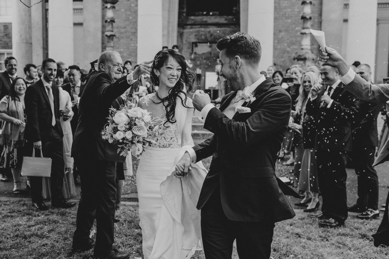 A happy couple exits their Wedding, showered with confetti. The bride, in a white gown, smiles at her groom in a dark suit, surrounded by joyous guests.