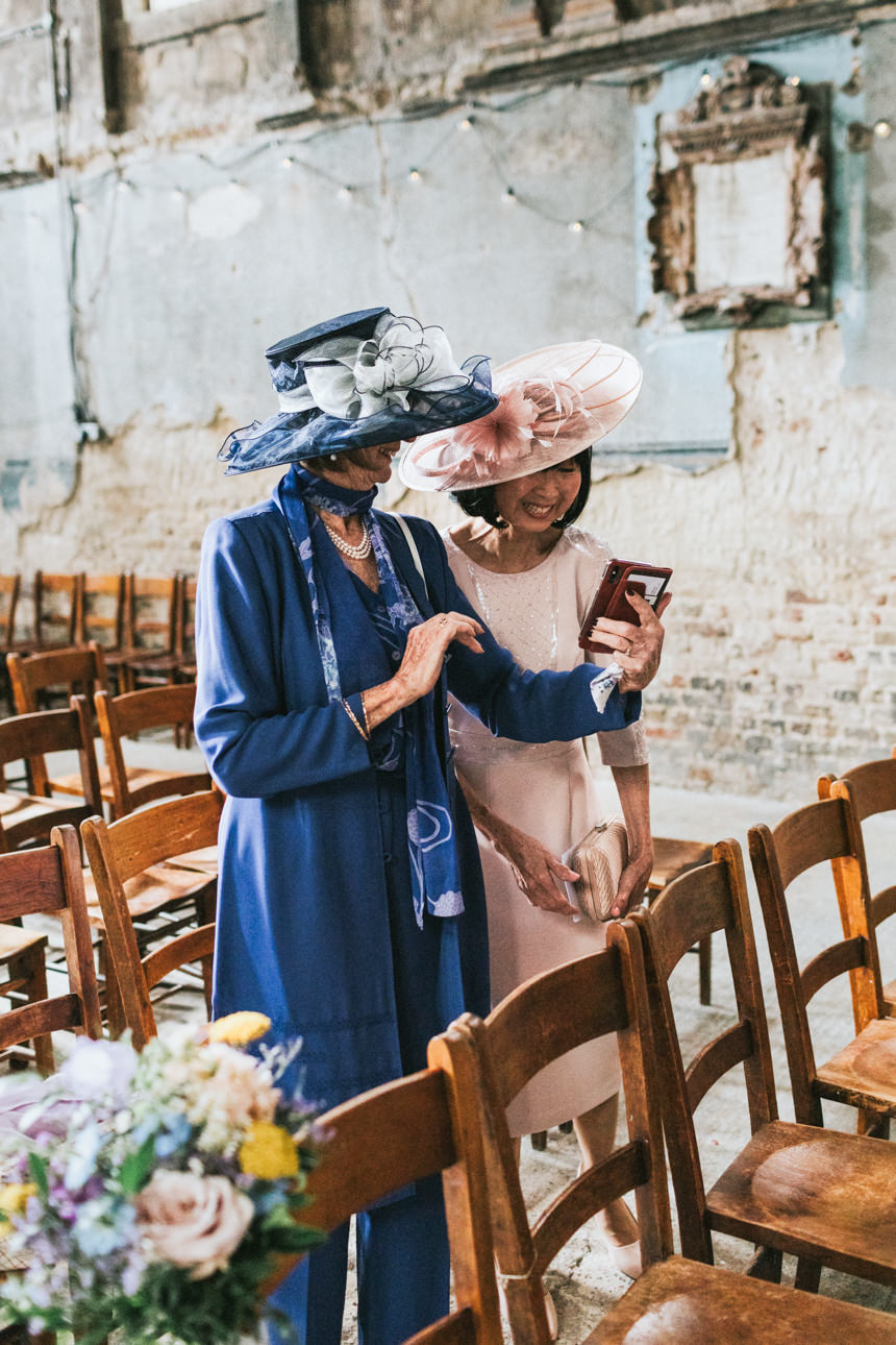 Two elegantly dressed women in hats look at a phone during a Brunswick House Wedding. One wears a blue suit and ornate hat, the other a pink dress and hat, amidst rows of wooden chairs.