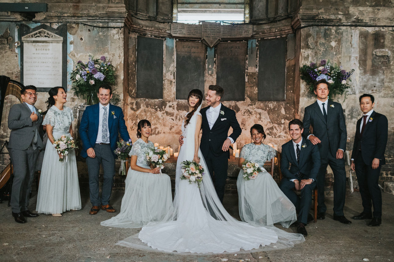Brunswick House and Asylum Chapel Wedding: Bride and groom pose with their wedding party in a historic venue. The bride wears a white gown, the groom a black suit, and the wedding party is in muted grey and blue tones.