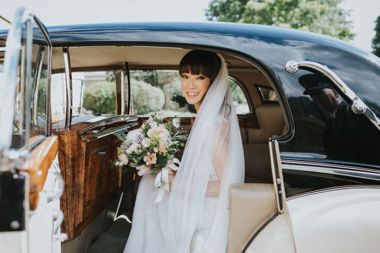 A radiant bride arrives at her Brunswick House Wedding, stepping out of a classic black and white car. She's holding a pastel bouquet and wearing a flowing white veil, smiling brightly.