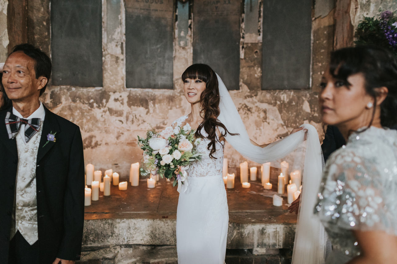 Brunswick House Wedding: A radiant bride in a white lace gown, holding a bouquet, stands near her father and a wedding guest amidst candlelight, creating a warm, intimate atmosphere.