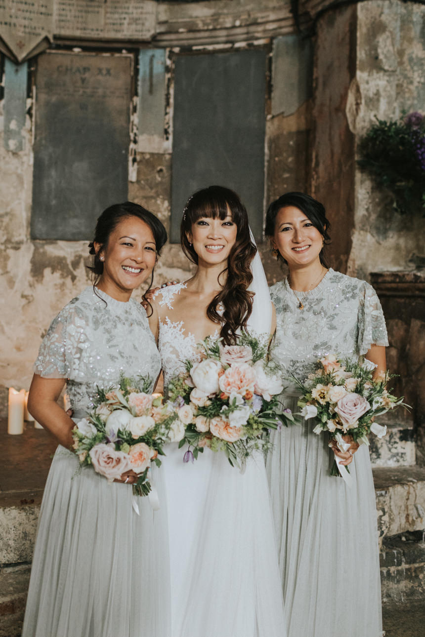 Bride and bridesmaids at a Brunswick House Wedding. The women smile holding bouquets of pink and peach flowers, wearing matching grey dresses with lace tops. Stone walls are in the background.