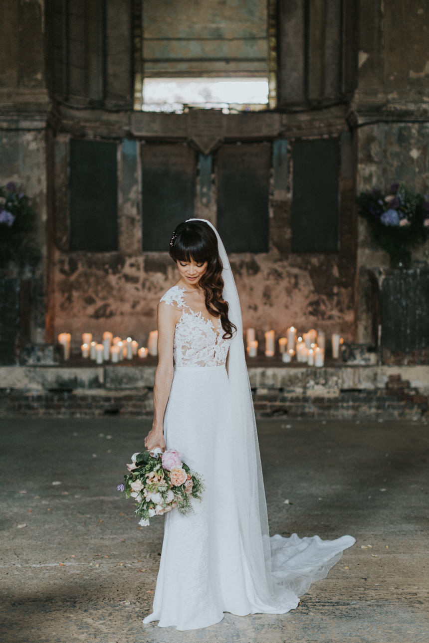 Brunswick House Wedding: Bride with cascading veil and lace dress holds a pastel bouquet, standing before a backdrop of candles and aged stone walls.