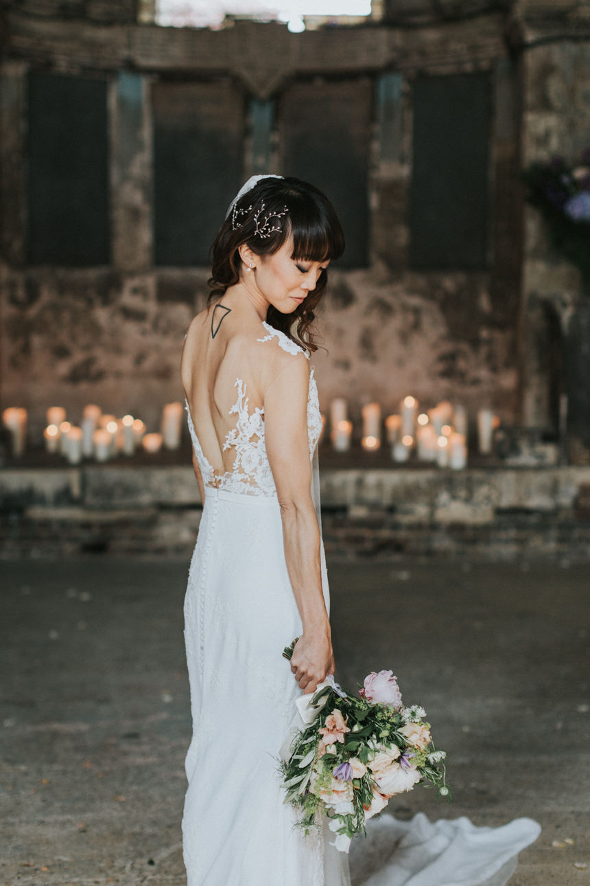 Brunswick House Wedding: Bride in a white lace gown holds a bouquet, back to the camera, with candles softly lighting the background.