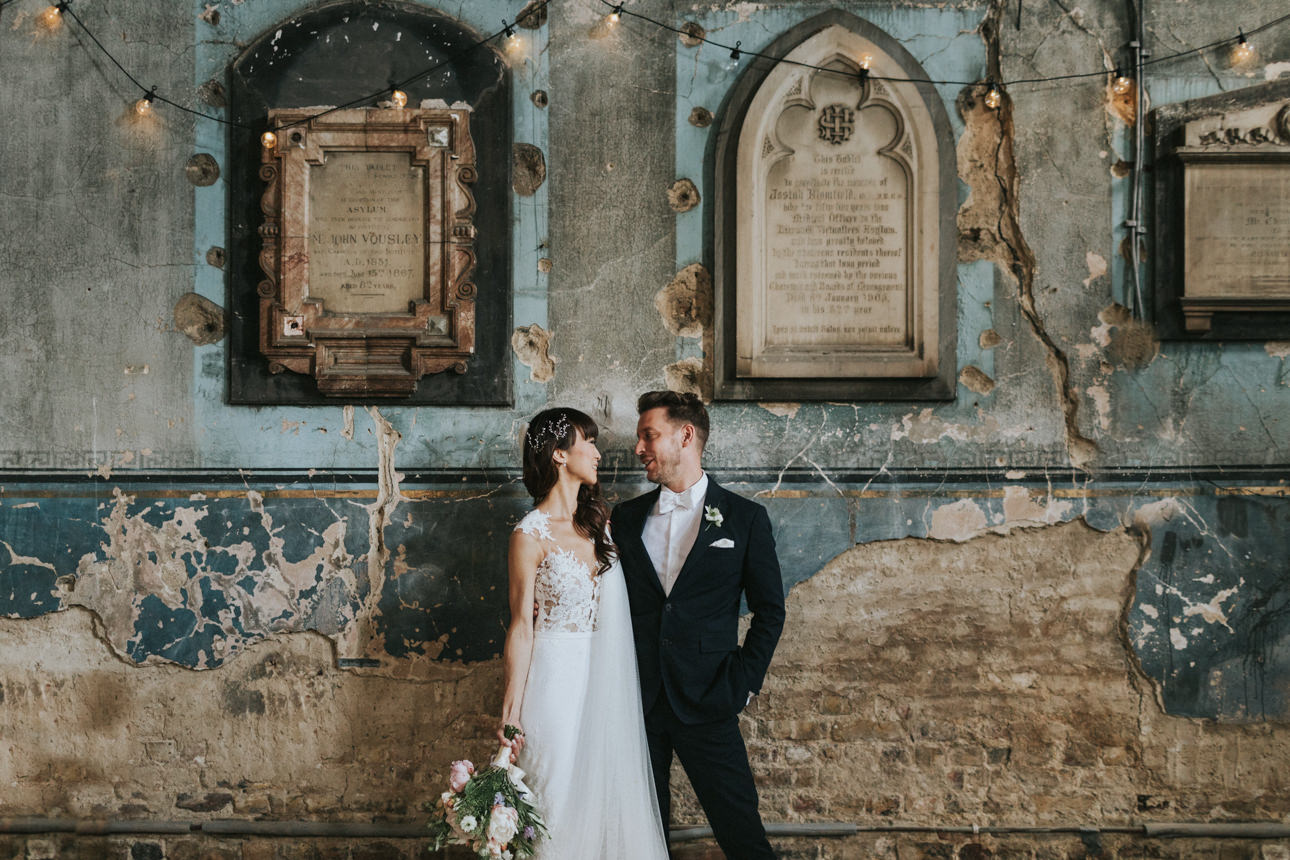 A stylish couple during their Brunswick House Wedding, gazing at each other in front of a distressed wall with aged plaques and string lights. The bride wears a lace dress, and the groom a sharp black suit.