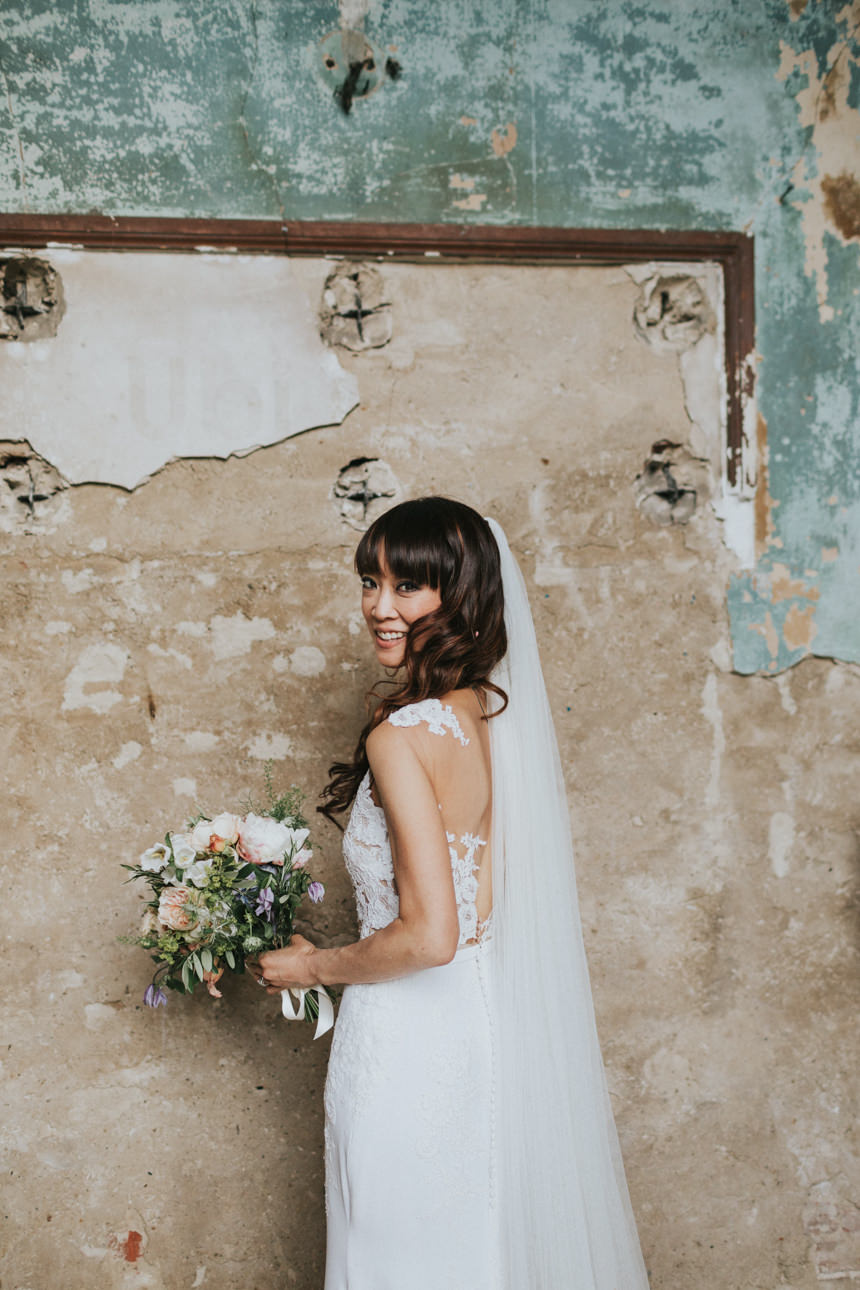 Bride smiles, holding a bouquet in her white lace gown and veil, against a textured, distressed wall with a framed section.