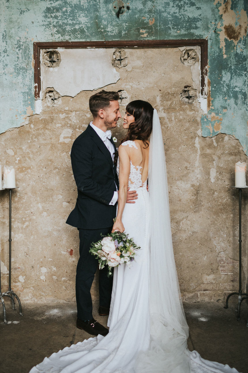 Brunswick House and Asylum Chapel Wedding: A bride and groom embrace in front of a weathered wall. The bride wears a white lace gown and long veil; the groom, a dark suit. Candles flank the loving couple.