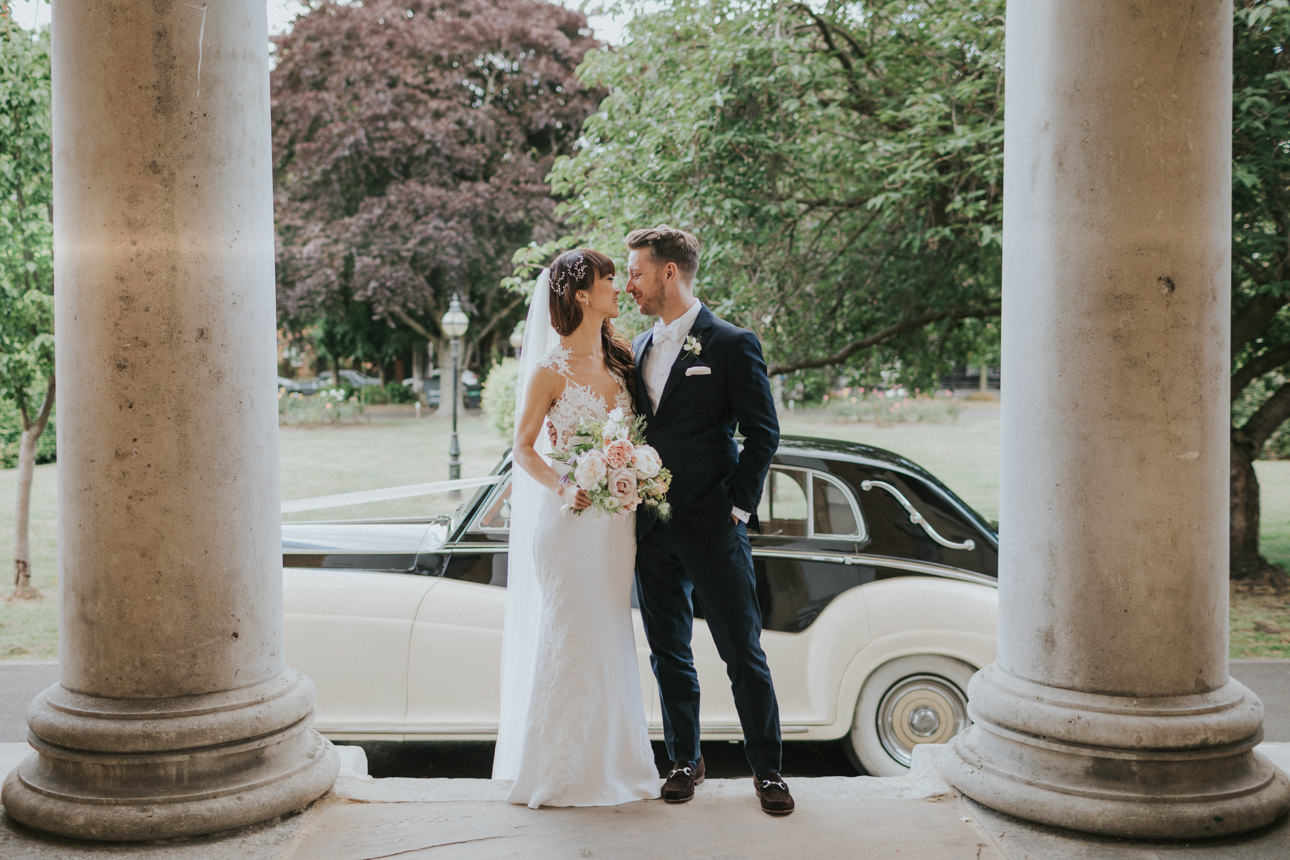 Bride and groom share a loving glance at their Brunswick House Wedding, framed by stone pillars and a vintage Rolls Royce. Her white lace gown complements his navy suit.