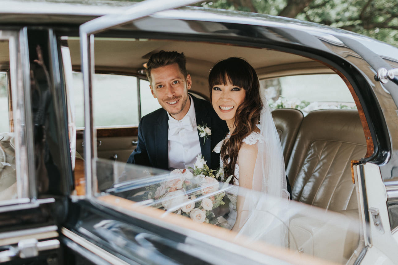 Newlyweds smile from inside a vintage car after their Brunswick House wedding. The bride, in a white dress and veil, holds a bouquet, while the groom wears a navy suit and bow tie.