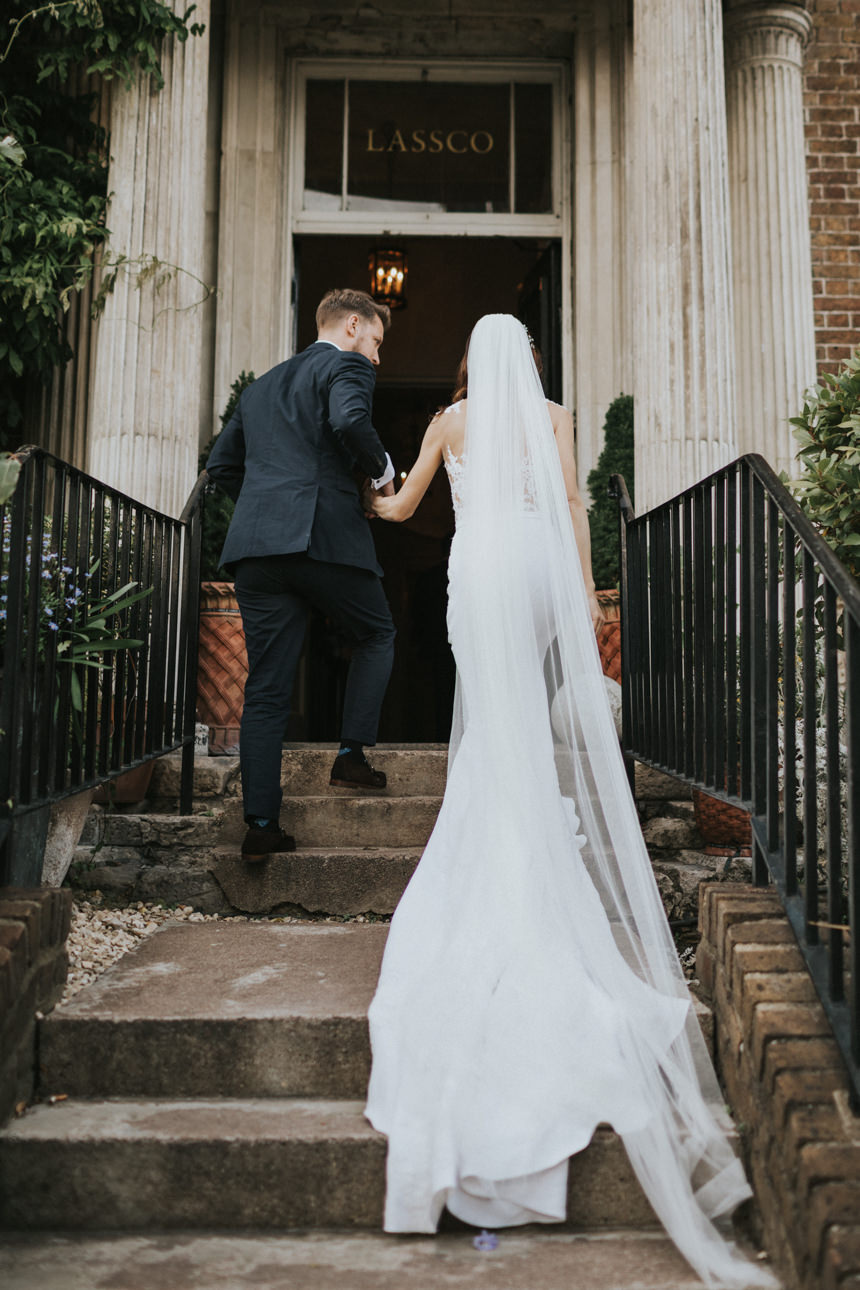 Bride and groom ascending steps to LASSCO Brunswick House after their Brunswick House wedding. The bride wears a flowing white gown and veil, and the groom wears a navy suit.