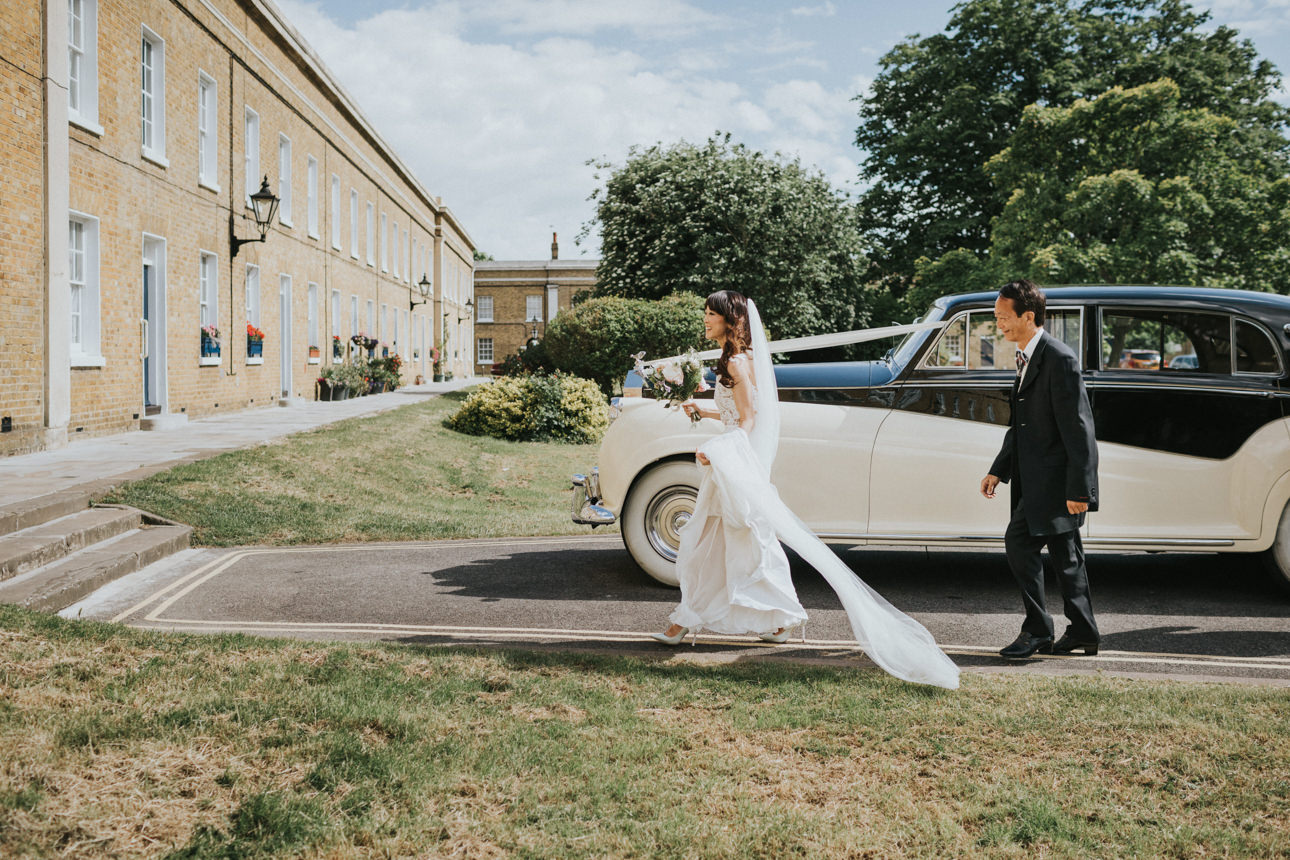 Bride in a white gown and veil walks with a man in a suit beside a vintage car, likely arriving at a Brunswick House Wedding. A brick building and greenery form the backdrop.