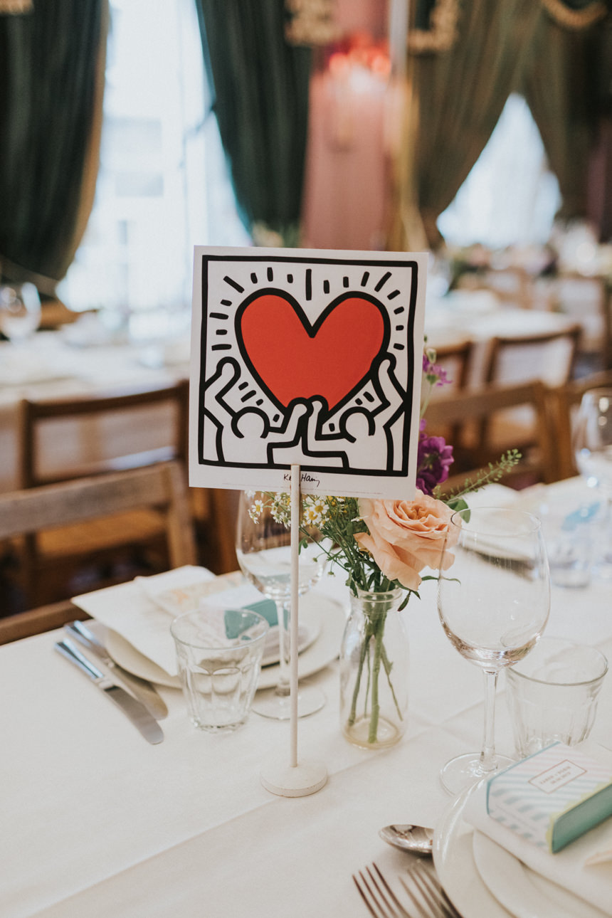 Table setting at a Brunswick House Wedding featuring a Keith Haring-inspired table number with a red heart, flowers, and glassware on a white tablecloth.