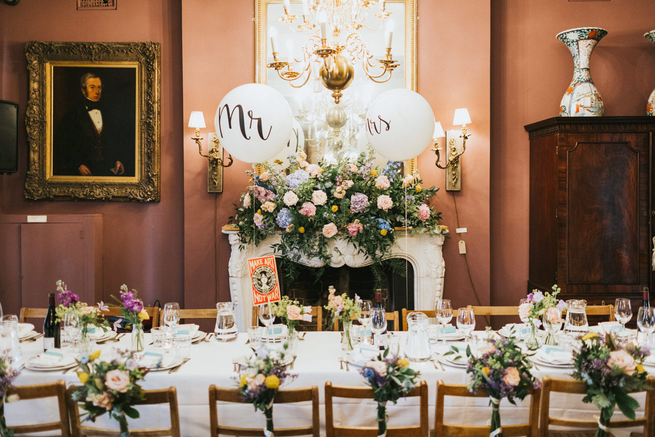 Brunswick House Wedding reception with a long table set for a feast. "Mr" and "Mrs" balloons float above a floral-covered mantelpiece. A formal portrait hangs on the mauve wall.