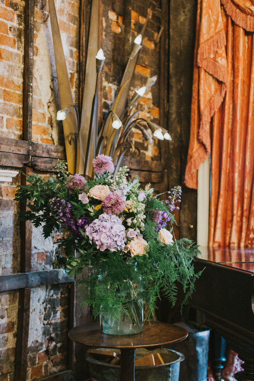 Floral centerpiece at a Brunswick House Wedding. Lush arrangement of pink, purple, and white flowers in a glass vase, set against a rustic brick wall and antique decor.