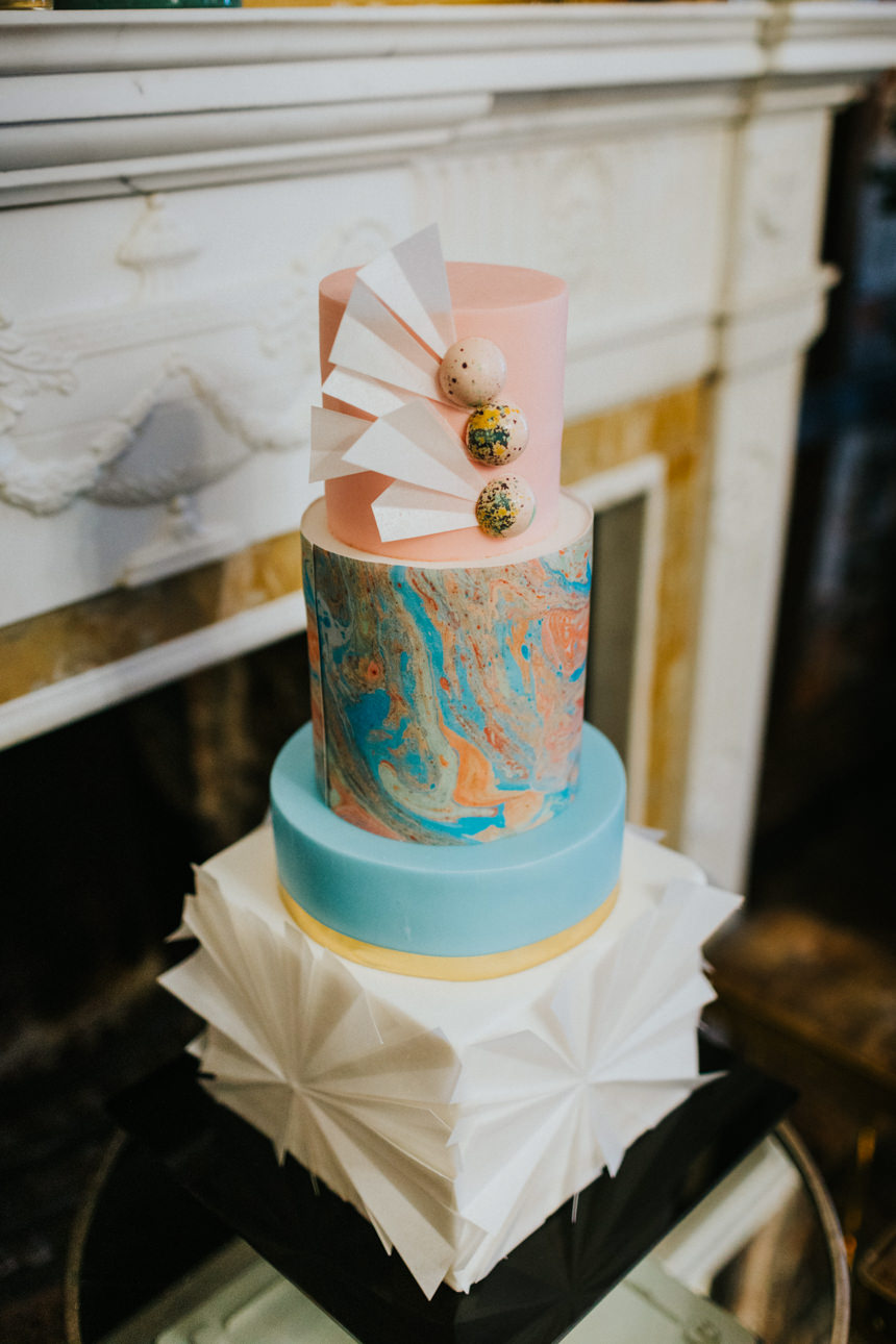 A modern, three-tiered cake at a Brunswick House Wedding. The tiers feature pink, marbled blue and orange, and white origami-like decorations. Decorated with white paper accents and speckled chocolate spheres.