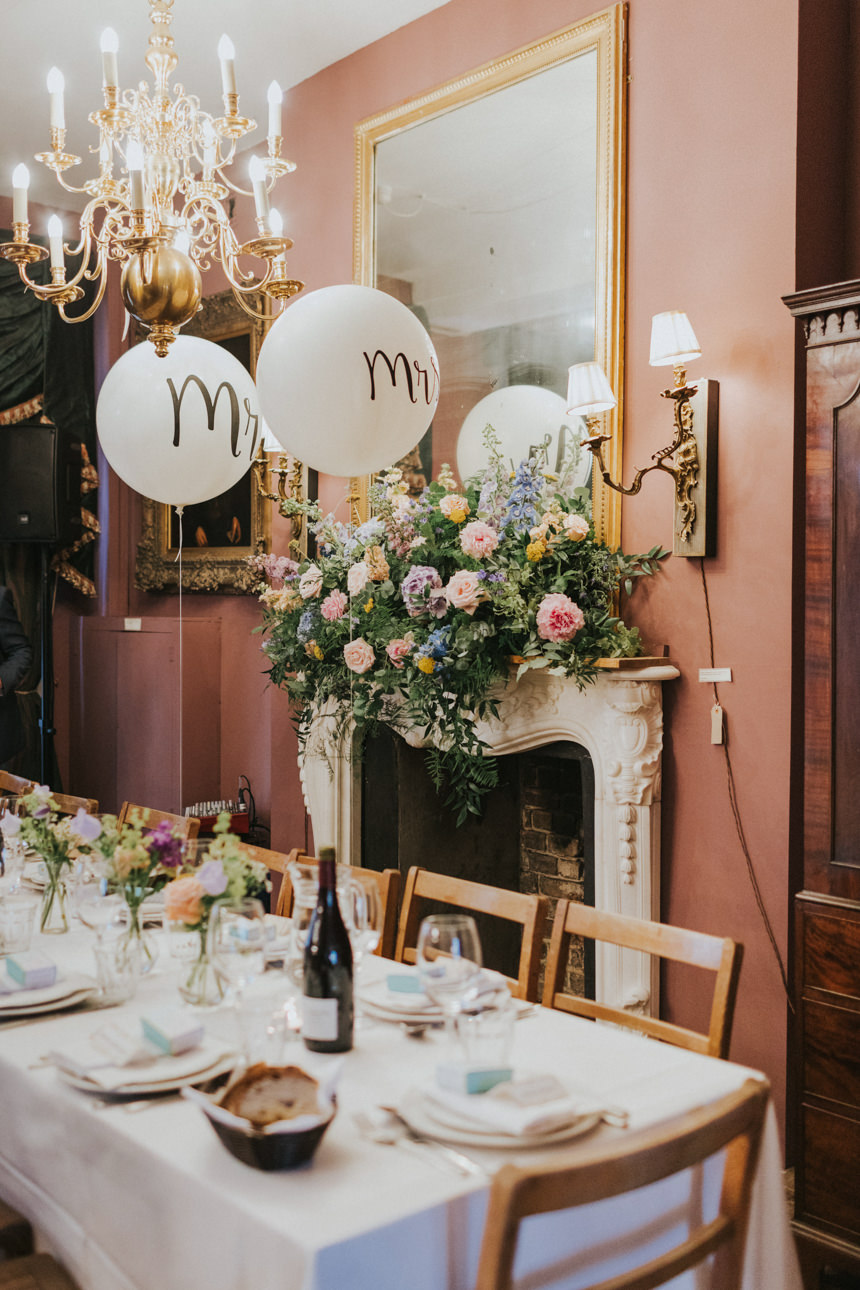 Brunswick House Wedding reception: Elegant table setting with white linens, floral centerpieces, and "Mr" balloons. Ornate chandelier and mantel with mirror.