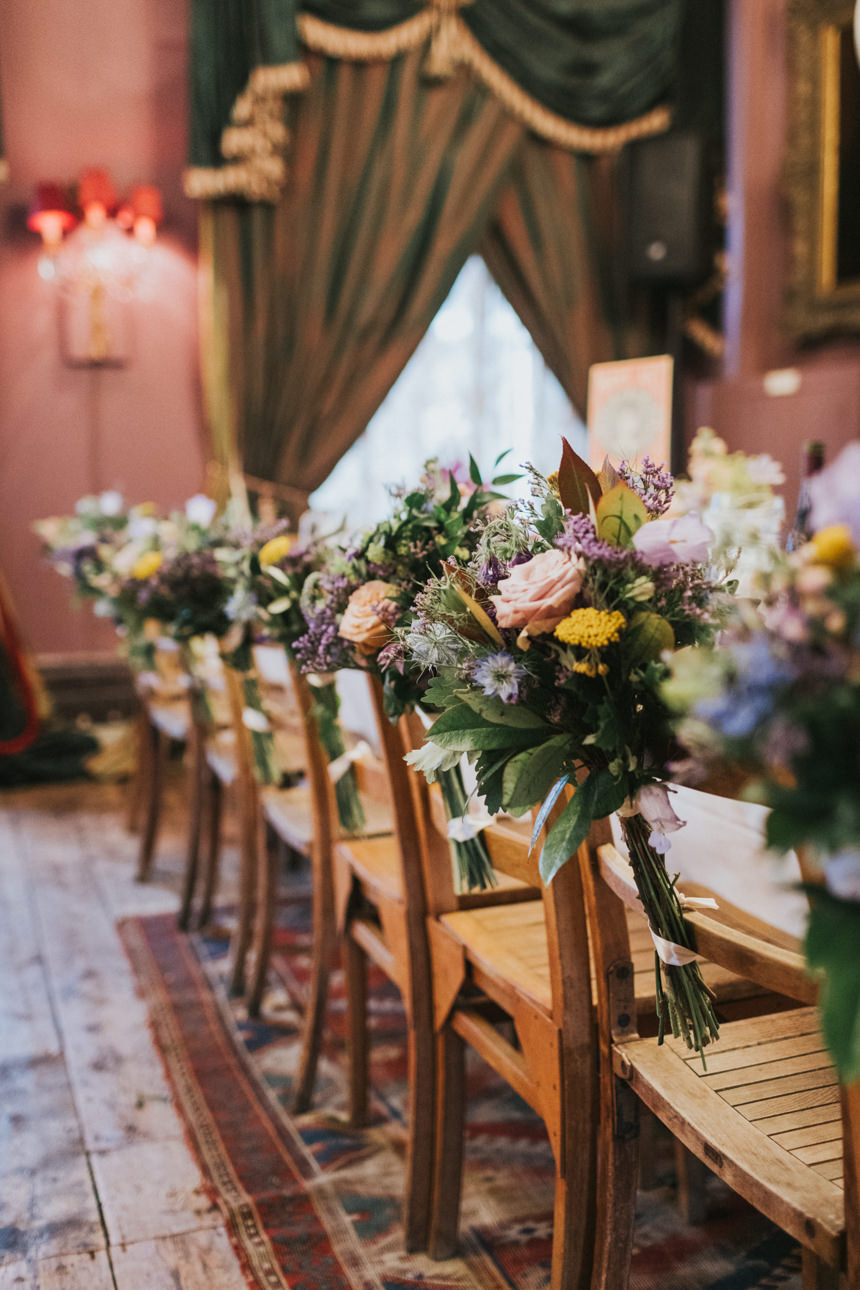 Wedding aisle decoration at a Brunswick House Wedding. Wooden chairs lined with colorful floral bouquets tied with ribbon, against a backdrop of ornate curtains and a vintage rug.