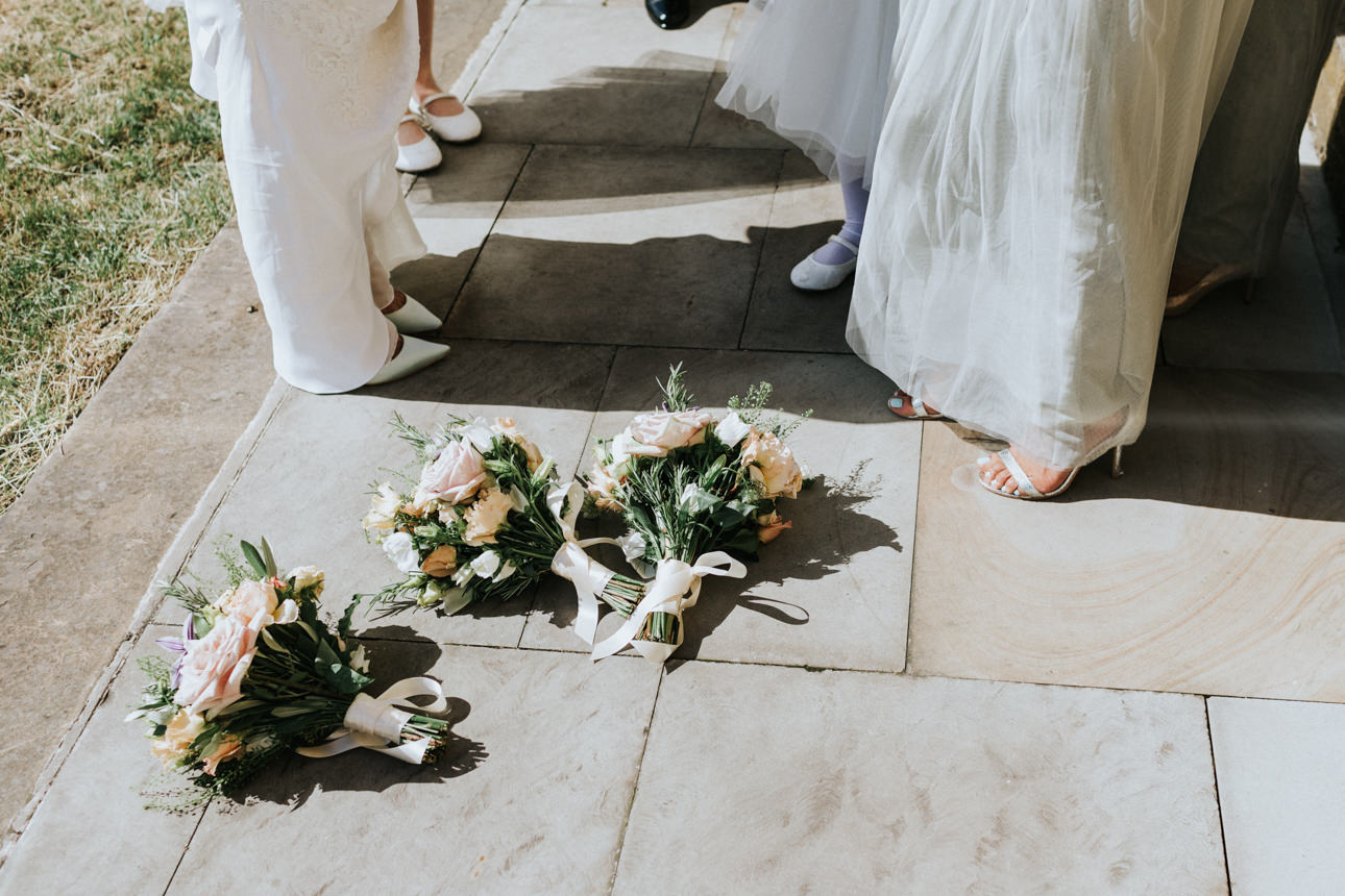Wedding bouquets rest on the stone patio at a Brunswick House Wedding. Soft pink and peach roses, tied with white ribbons, sit near the feet of wedding guests in white dresses and shoes.