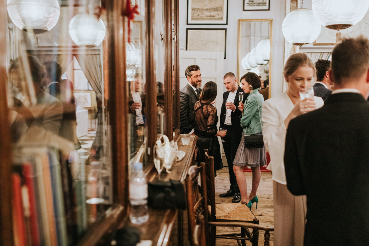 Guests mingle during a Brunswick House Wedding reception. A group of stylishly dressed attendees chat and hold champagne glasses in an elegantly decorated room with vintage furniture and globe pendant lights.
