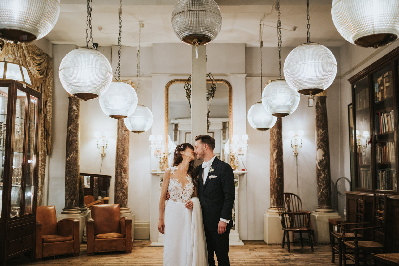 A couple shares a kiss at their Brunswick House wedding. The bride wears a white dress with lace detailing, while the groom is in a black suit. Ornate chandeliers and antique decor fill the room.