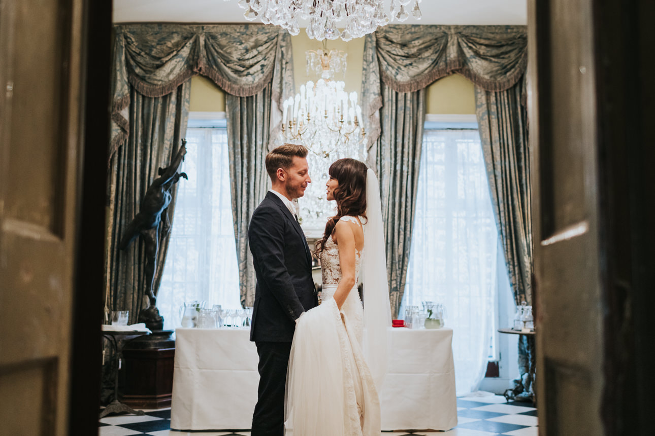 Bride and groom share a moment at their Brunswick House Wedding, framed by ornate drapes and a chandelier. Black and white checkered floor.