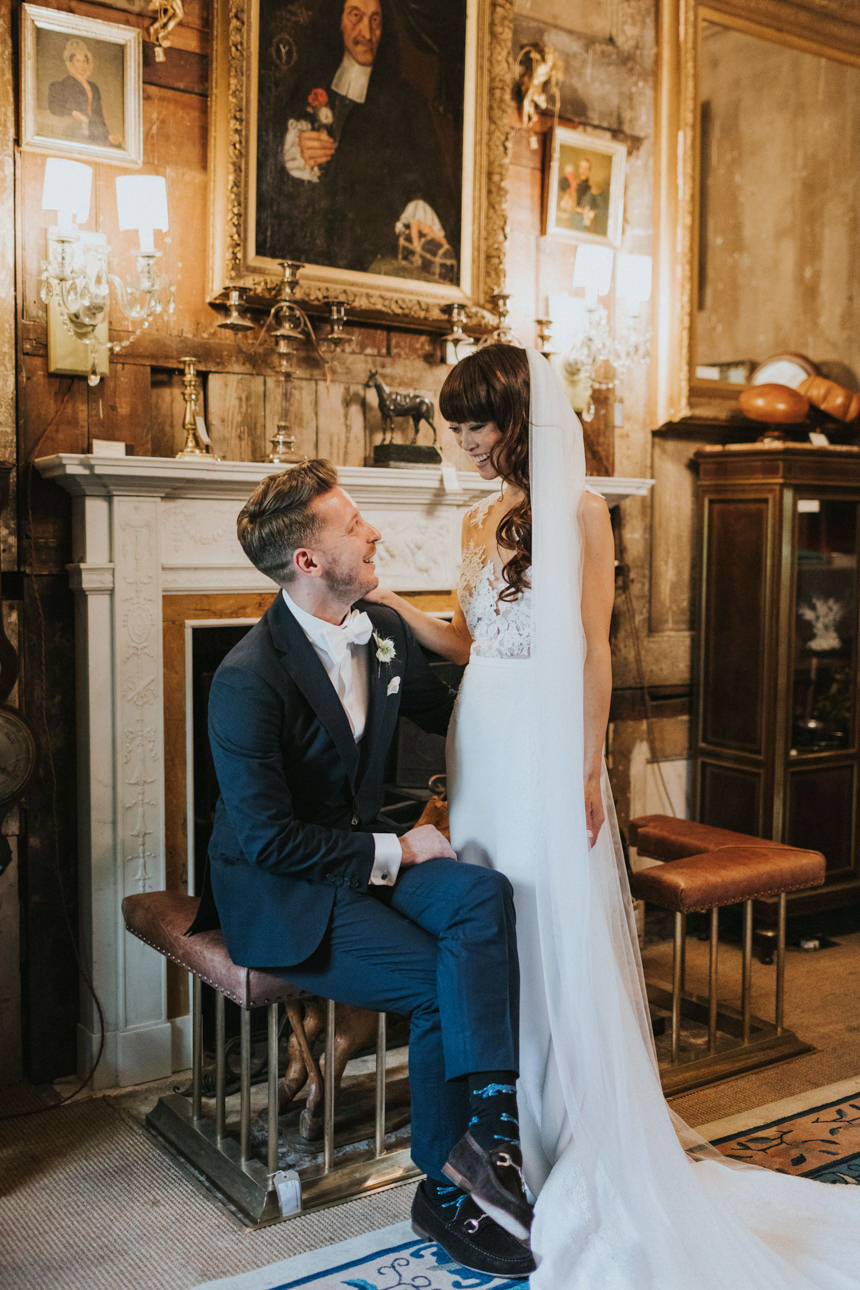 Brunswick House Wedding: Bride and groom share a loving glance. She wears a lace dress and long veil, he's in a navy suit with fun socks, set in an ornate room with paintings and a fireplace.