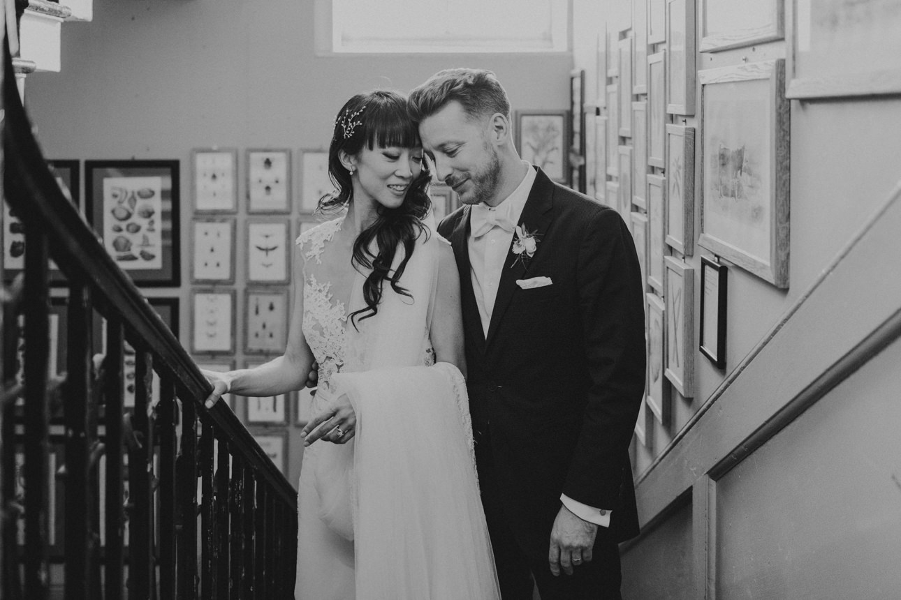 A bride and groom share a tender moment on the stairs at their Brunswick House Wedding. The bride, with dark hair and bangs, wears a lace dress, while the groom sports a dark suit and bow tie.