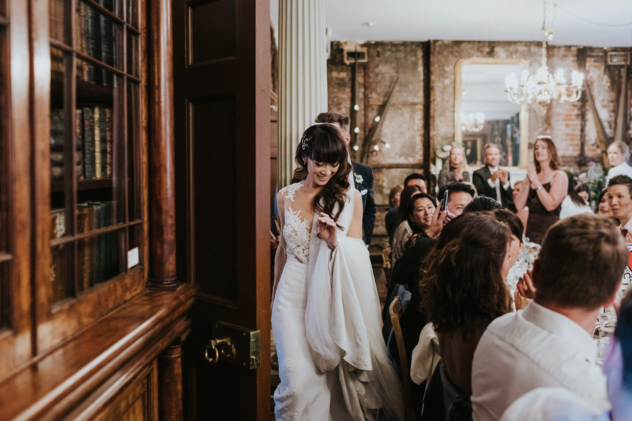 A radiant bride enters Brunswick House for her wedding reception, her lace gown and flowing train catching the light. Guests look on with smiles and applause in the warmly lit, antique-filled room.
