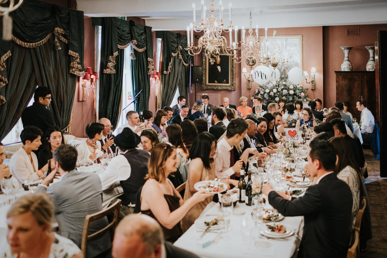 Brunswick House Wedding: Guests seated at a long, elegantly set table in a grand, candlelit room, enjoying a meal and conversation. Dark green curtains and chandeliers add to the sophisticated ambiance.