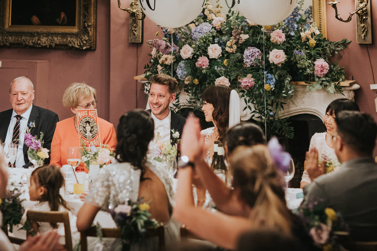 Brunswick House Wedding: A joyful wedding reception scene with the bride and groom smiling at the head table, surrounded by guests and a lush floral arrangement. Guests are clapping and celebrating.