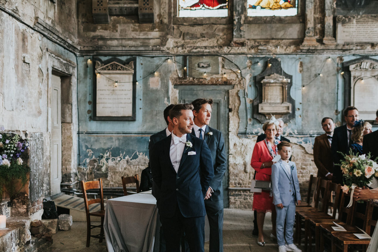 Two grooms await their Wedding ceremony in a weathered, historic room with peeling blue paint and stained glass. Guests, including a young boy in a light blue suit, look on.