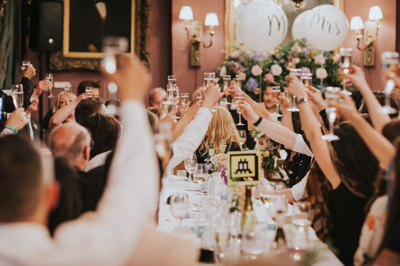A jubilant Brunswick House Wedding scene: Guests raise champagne glasses in a toast at a long, elegantly set table, celebrating the newlyweds amidst soft lighting and floral decorations. "Mr" and "Mrs" balloons float in the background.