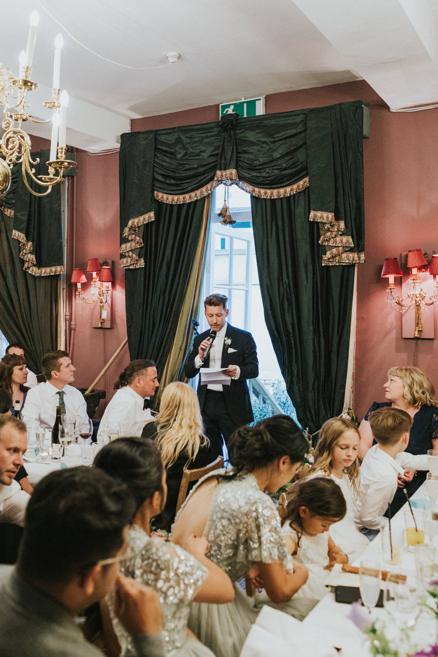 A groom gives a speech at his Brunswick House Wedding, standing before dark green drapes. Guests sit at tables, listening attentively in the warmly lit room.