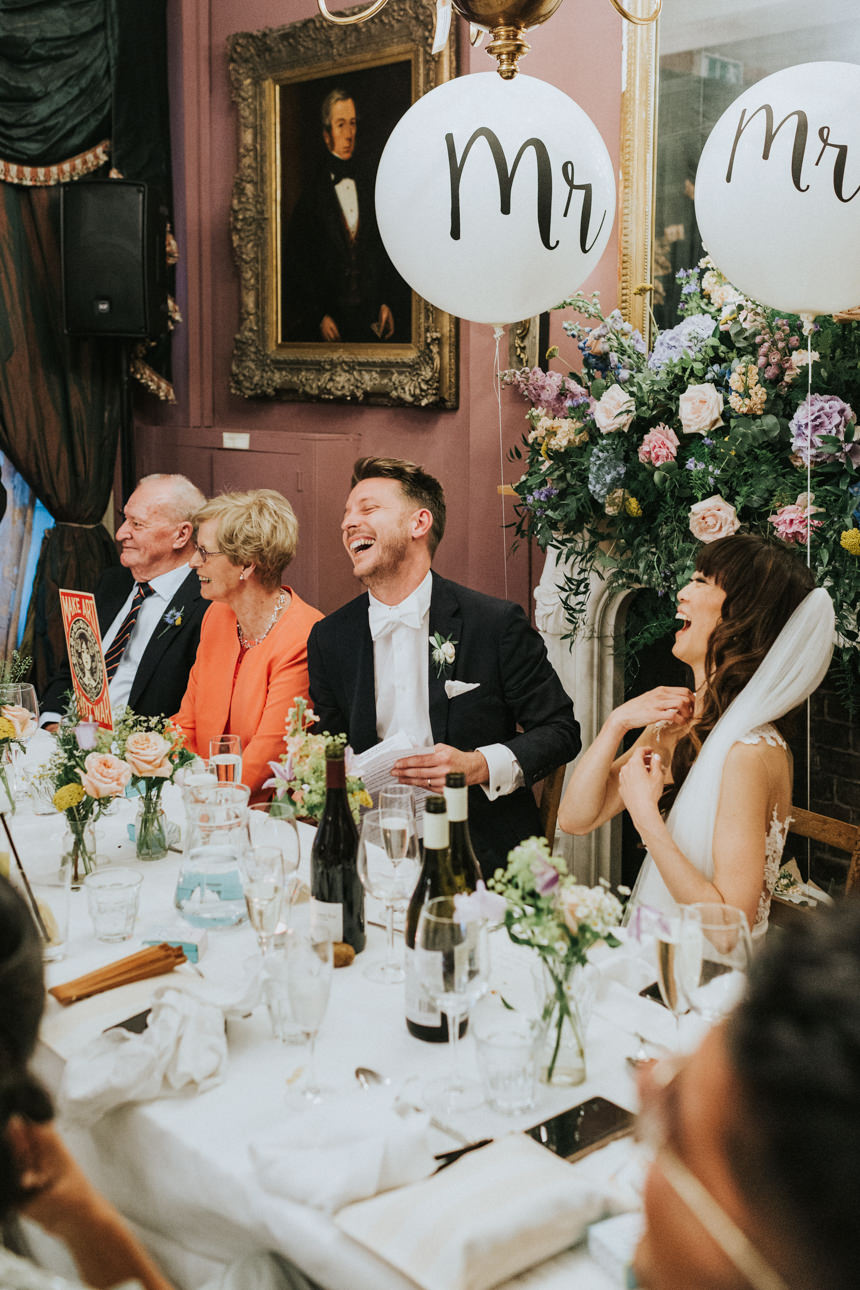 A joyous Brunswick House Wedding scene: the bride and groom share a laugh at the head table, decorated with flowers and "Mr" balloons, surrounded by family.