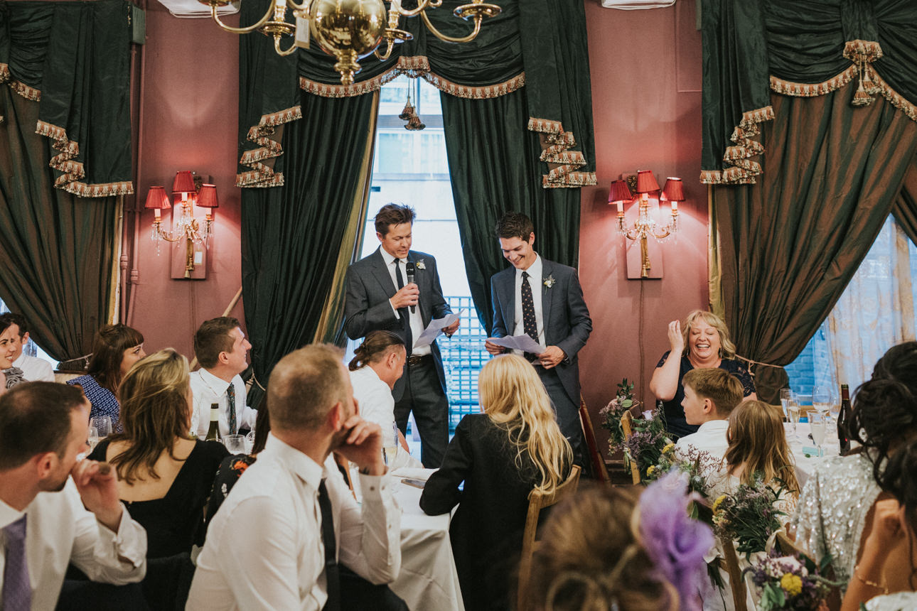 Two men giving a speech at a Brunswick House wedding reception. Guests are seated at tables, listening and laughing. The room has dark green drapes and red sconces.