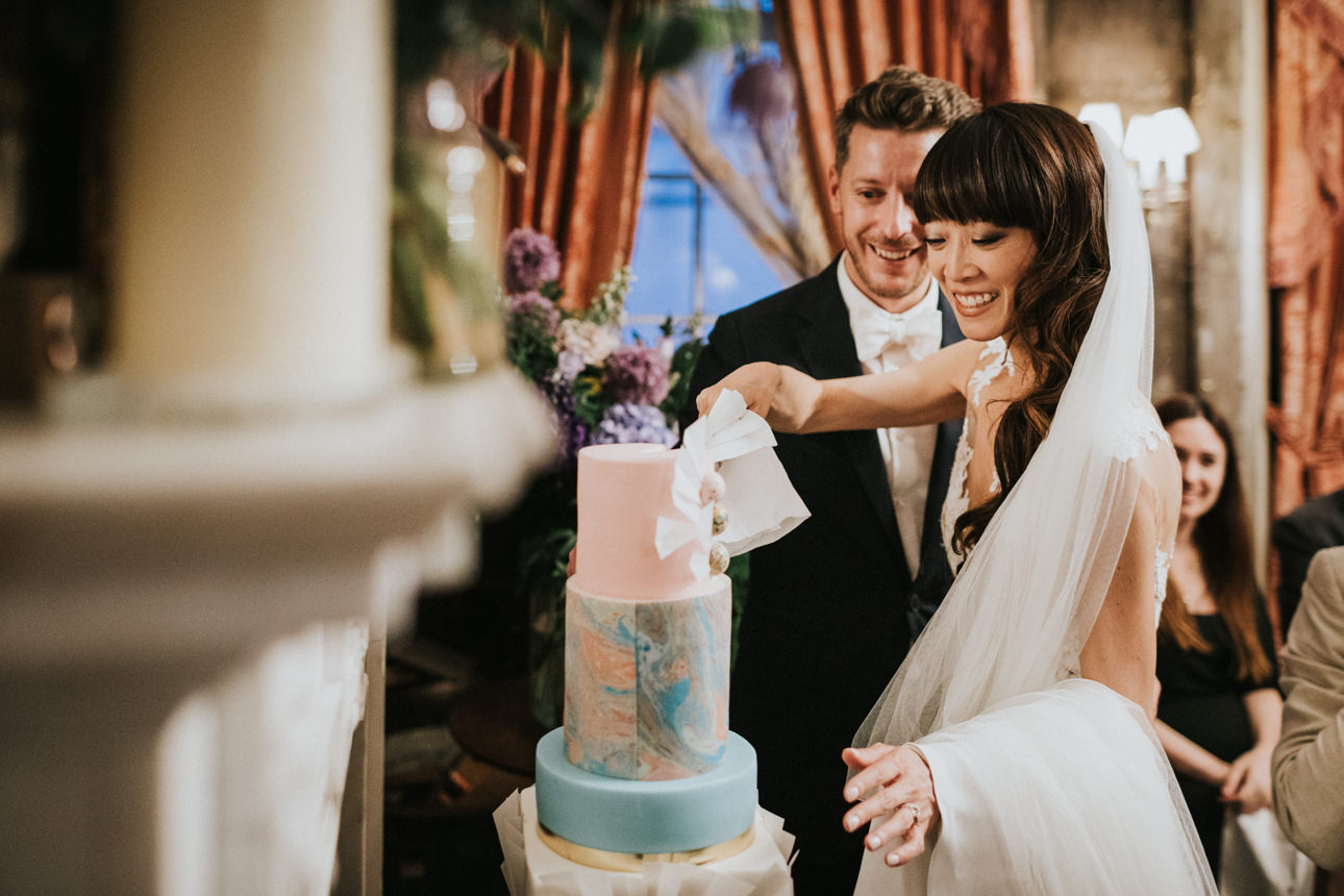 A smiling bride and groom cut their wedding cake at their Brunswick House Wedding. The three-tiered cake has pastel pink, blue, and marbled layers.
