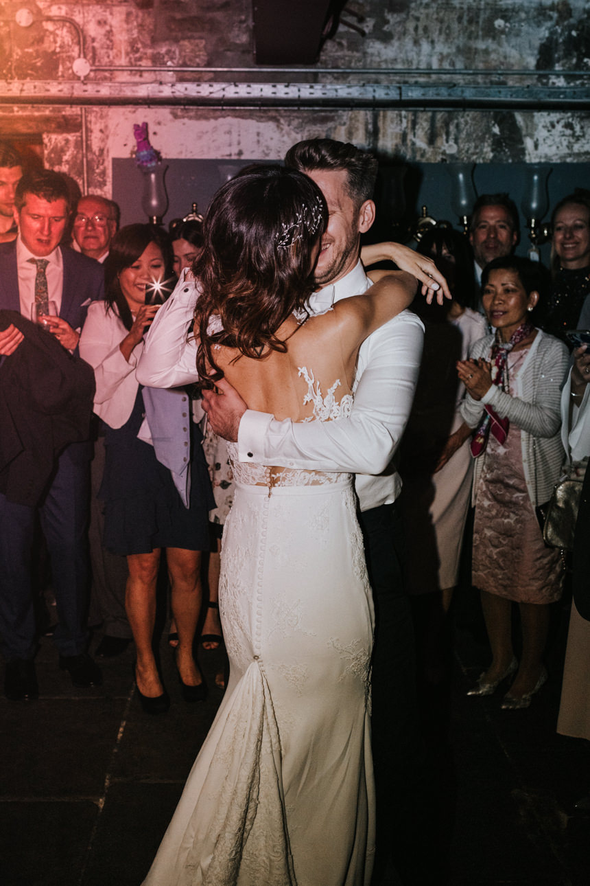 Bride and groom embrace during their first dance at a Brunswick House Wedding, surrounded by cheering guests. The bride wears a white lace dress with a train.