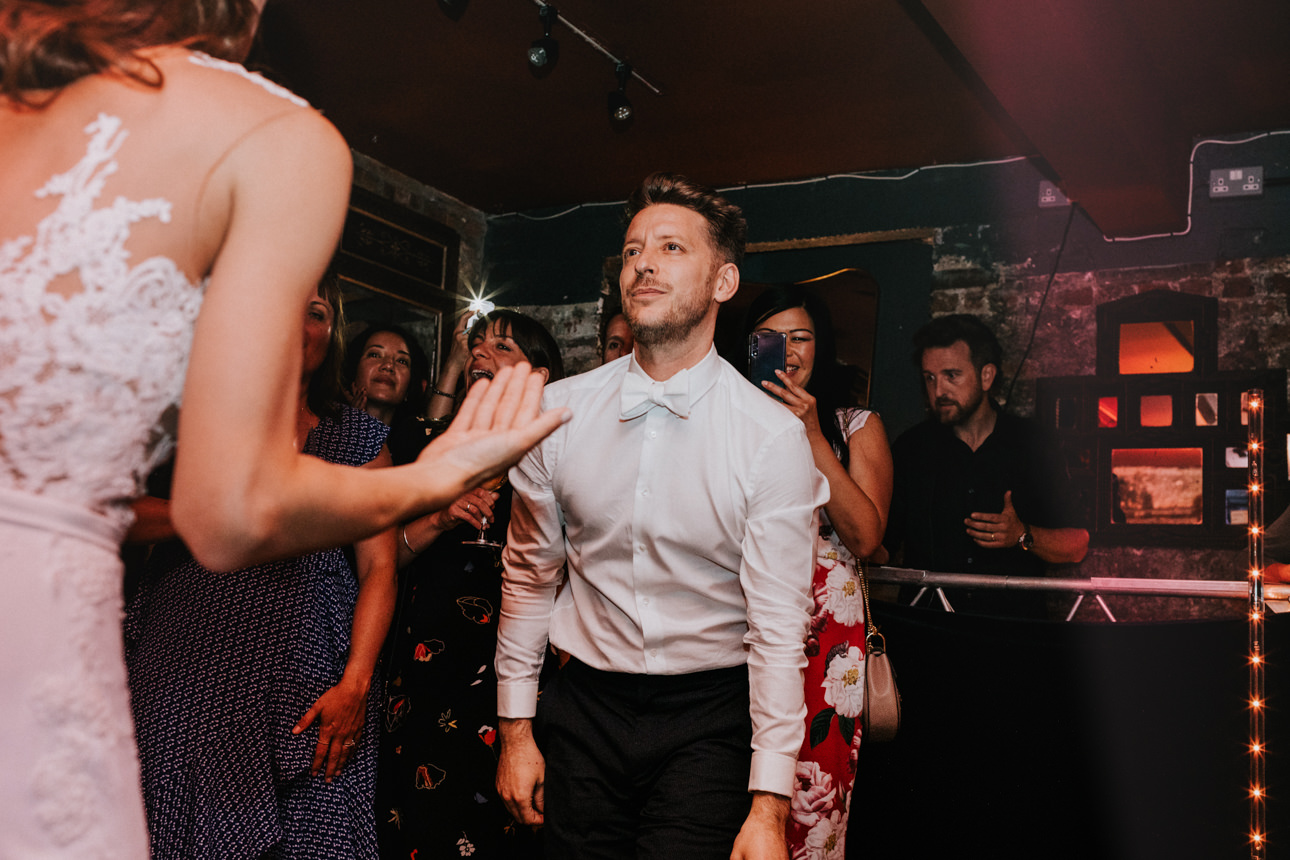 Groom on the dance floor at a Brunswick House Wedding, eyes up, surrounded by guests in a dimly lit room. A bride in white is partially visible on the left.