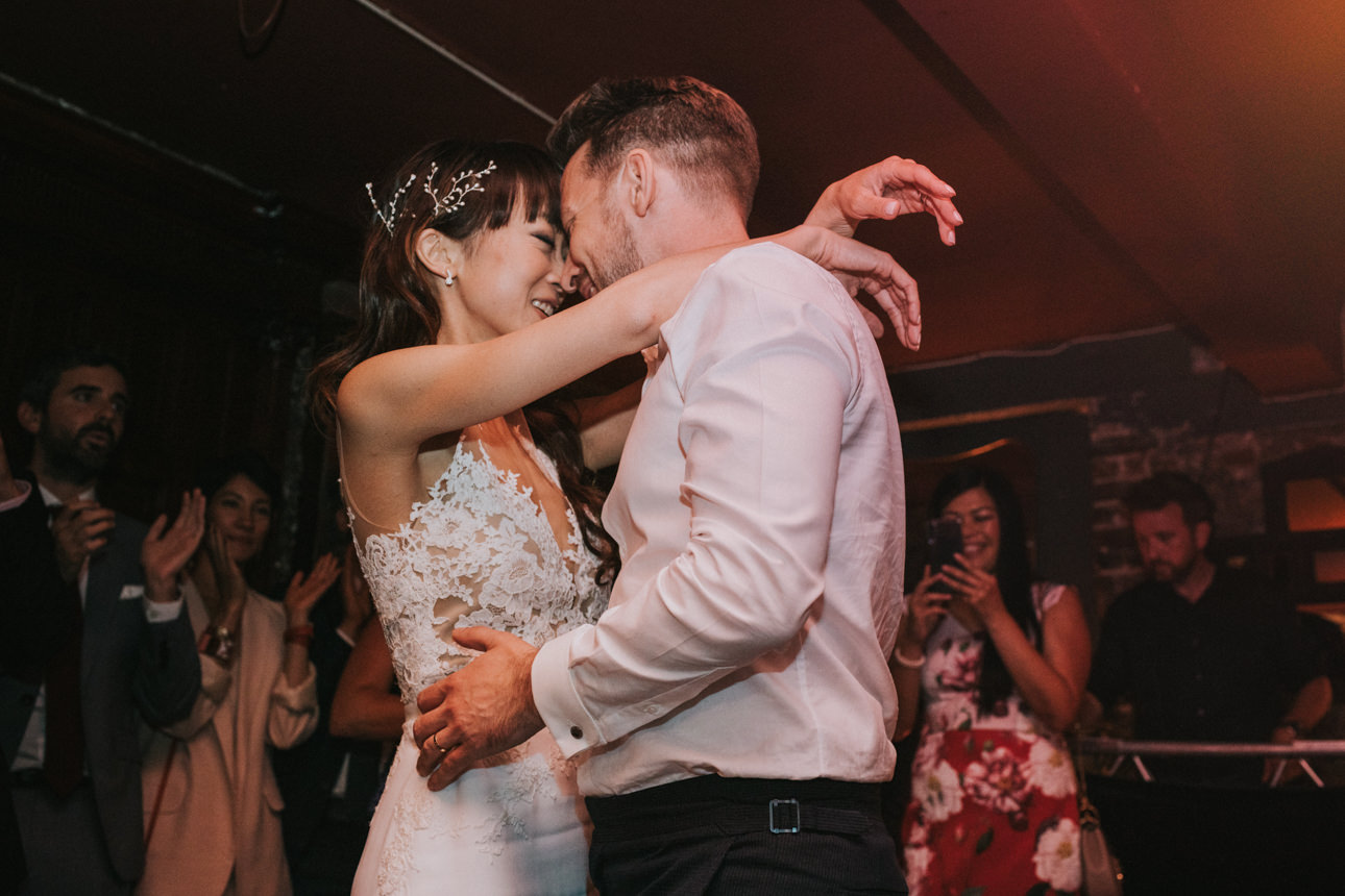 Bride and groom embrace during their first dance at their Brunswick House Wedding. The bride wears a white lace dress and the groom a white shirt. Guests look on in the background.