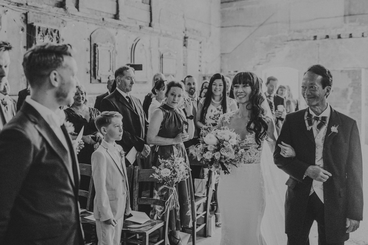 Bride walks down the aisle at her Brunswick House and Asylum Chapel Wedding, arm-in-arm with her father. Guests watch with smiles in the unique, rustic venue.