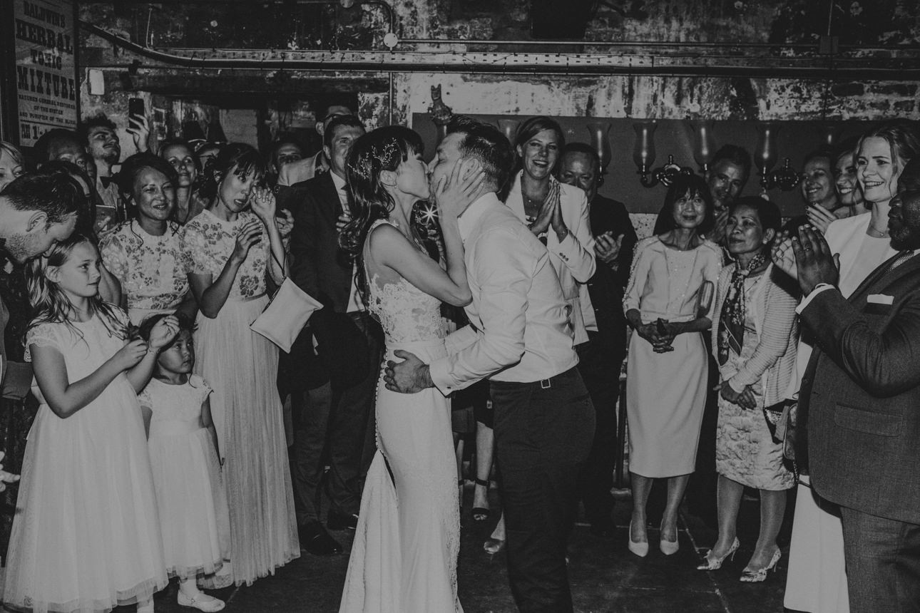 A bride and groom share a kiss surrounded by wedding guests. The bride wears a white lace gown, while the groom is in a white shirt and dark pants. Guests clap and cheer in this candid moment.